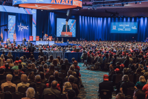 Eine große Menschenmenge sitzt auf Stühlen, die einer Bühne zugewandt sind, mit einigen Personen, die auf dem Podium und auf der linken Seite stehen, blaue Vorhänge und ein Bildschirm im Hintergrund und ein orangefarbenes Schild oben, das das CPAC 2020-Event am 25. Oktober anzeigt.