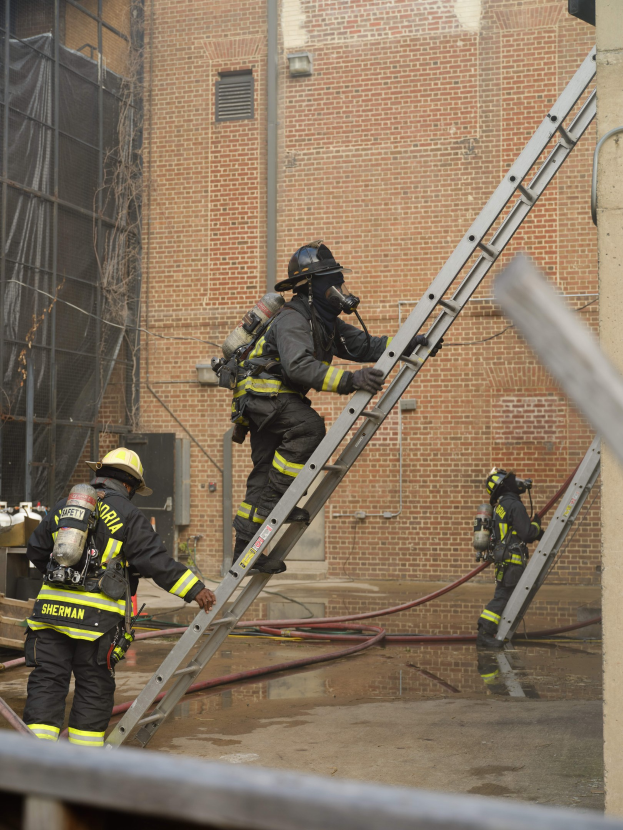 Feuerwehrleute in Helmen und Ausrüstung, die eine Leiter vor einem Backsteingebäude hochklettern, mit Rohren am Boden und einer Metallstange unten, sowie einem weiteren Gebäude mit Fenstern und einem Netz im Hintergrund.