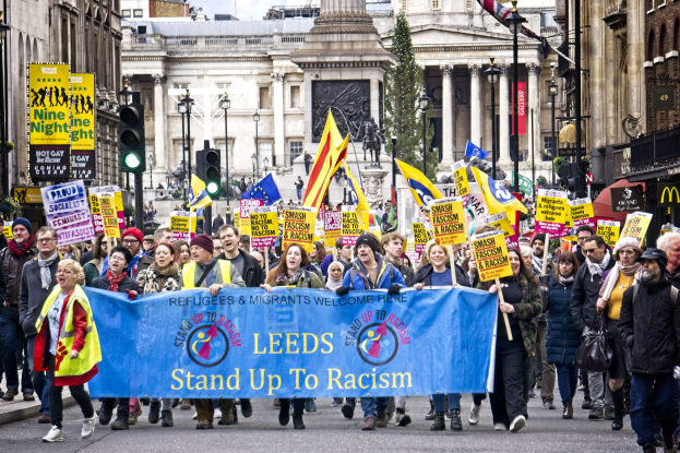 Eine Gruppe von Menschen marschiert mit einer 'Stand Up to Racism'-Schlagzeile die Straße entlang, mit Gebäuden, Bäumen, Laternen, Verkehrsampeln und einer Statue im Hintergrund bei bewölktem Himmel.