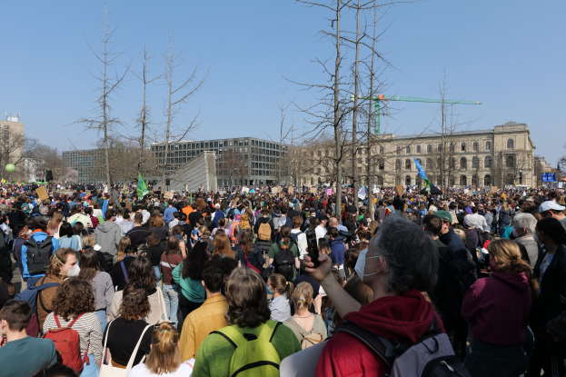 Eine große Menge Menschen mit Schildern und Töten vor einem Gebäude mit Fenstern, Bäumen und einem klaren Himmel im Hintergrund, was auf eine Klimaprotest Demonstration hinweist.