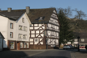 Holzhaus in einer Kleinstadt umgeben von Gebäuden, Fahrzeugen, Fußgängern, Bäumen und Straßeninfrastruktur unter einem klaren blauen Himmel.