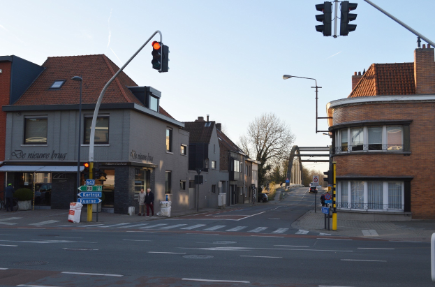 Stadtstraßenkreuzung mit Ampel, Fußgängern, Fahrrädern, Gebäuden, Bäumen und einem Tor unter einem klaren blauen Himmel.