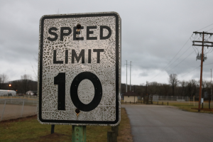 Ein Tempolimit-Schild am Straßenrand, umgeben von Gras, Strommasten mit Drähten, einem Zaun, Häusern, Bäumen und einem Himmel mit Wolken im Hintergrund.