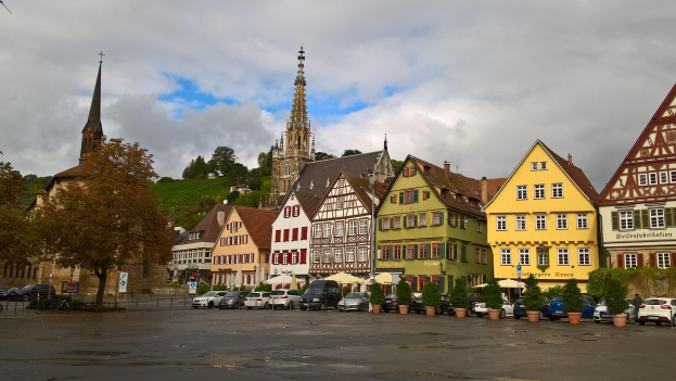 Ein malerischer Marktplatz in Rothenburg ob der Tauber, Deutschland, mit verschiedenen Gebäuden mit Fenstern, Bäumen, Topfpflanzen, Fahrzeugen, Schildern und einer Kirche im Hintergrund bei einem bewölkten Himmel.
