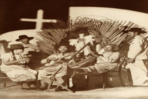 Schwarz-weiß-Foto einer Mariachi-Band, die vor einer Kirche mit einem Kreuz an der Wand dahinter sitzt.