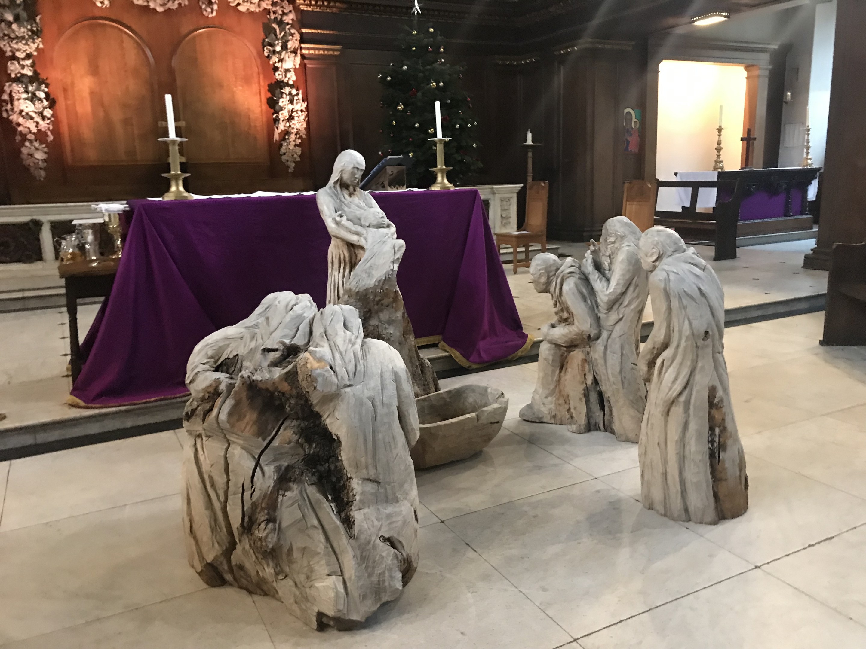 Eine Krippe in einer Kirche mit Statuen auf dem Boden, einem Tisch mit einer Decke und Kerzenständern, Stühlen, einem geschmückten Weihnachtsbaum und einer Wand im Hintergrund.