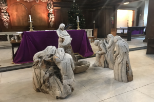 Eine Krippe in einer Kirche mit Statuen auf dem Boden, einem Tisch mit einer Decke und Kerzenständern, Stühlen, einem geschmückten Weihnachtsbaum und einer Wand im Hintergrund.