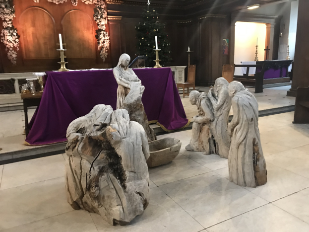 Eine Krippe in einer Kirche mit Statuen auf dem Boden, einem Tisch mit einer Decke und Kerzenständern, Stühlen, einem geschmückten Weihnachtsbaum und einer Wand im Hintergrund.
