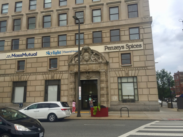 Großes Gebäude mit "Massachusetts Mutual Financial Group"-Schriftzug, parkende Autos, Straßenlaternen, fahrende Autos, Fußgänger auf dem Gehweg, Grünfläche und bewölkter Himmel.