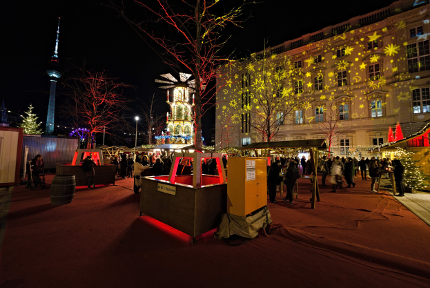 Ein belebter Weihnachtsmarkt in Berlin, Deutschland, mit Menschen um dekorierte Stände, festliche Lichter, Bäume, Gebäude, Laternenmasten und einen Turm unter einem dunklen Himmel.