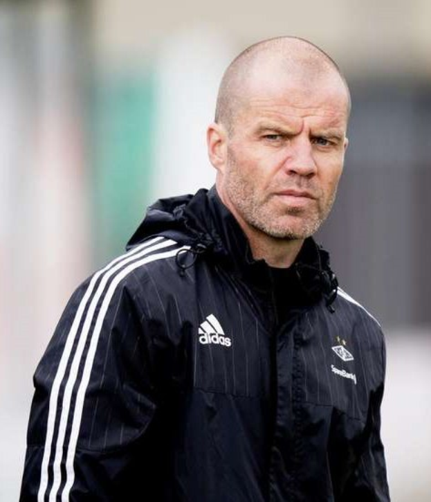 Ein kahlköpfiger Trainer in einer blauen Jacke mit weißen Streifen und weißem Kragen steht auf einem Fußballfeld mit verschränkten Händen und ernster Miene, unscharfer Hintergrund.