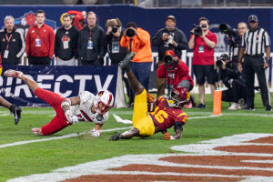 Ein Fußballspieler taucht in die Endzone ein, um einen Touchdown zu erzielen, mit einer Gruppe von Menschen am Boden, einige halten Kameras, und ein Banner mit Text im Vordergrund und Geländer, Pfosten und andere Gegenstände im Hintergrund.