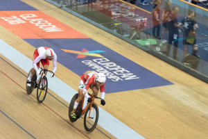 Zwei Männer mit Helmen fahren Fahrräder auf einer Bahn vor einem Publikum, rechts eine Glaswand mit der Aufschrift 'Europameisterschaften Glasgow 2018' und Menschen davor stehend und sitzend.
