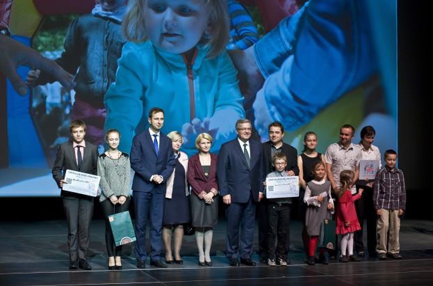 Eine Gruppe von Menschen auf einer Bühne mit Taschen und Urkunden, im Hintergrund ein Bildschirm mit einem Kind und anderen Personen.