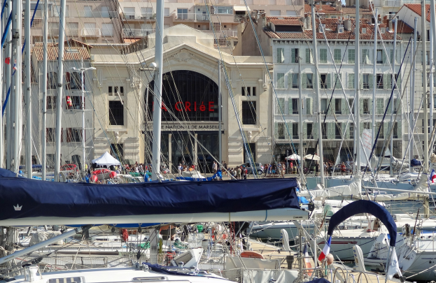 Ein Hafen mit zahlreichen vertäuten Booten, Gebäude mit Fenstern und einem Namensschild im Hintergrund sowie Zelte mit anwesenden Menschen.