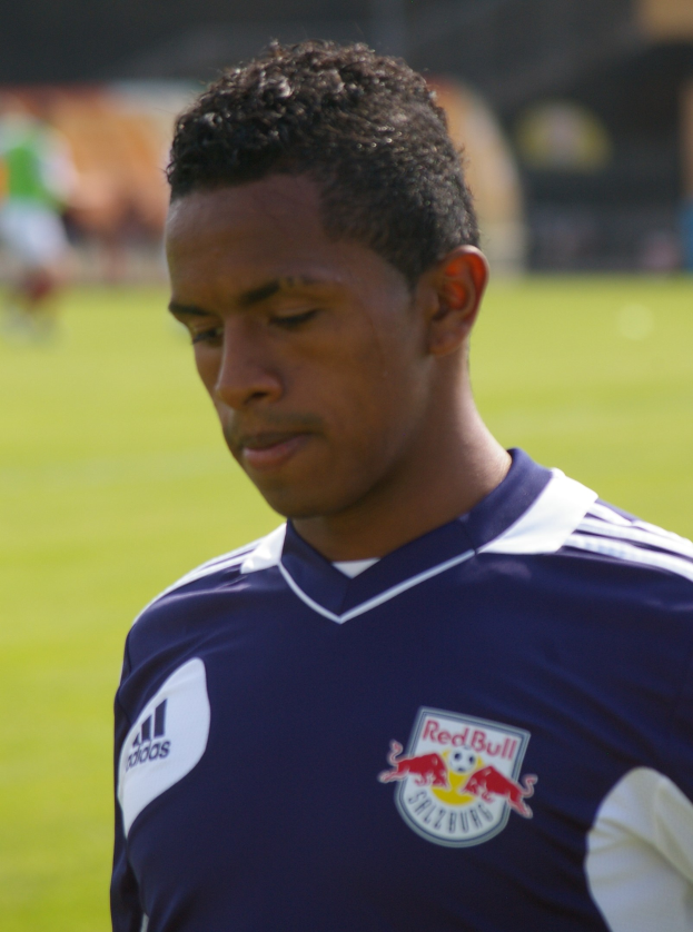 Junger Mann in blauem T-Shirt auf einem Fussballfeld, der wie ein Flügelspieler mit kurzem dunklem Haar und entschlossener Miene aussieht.