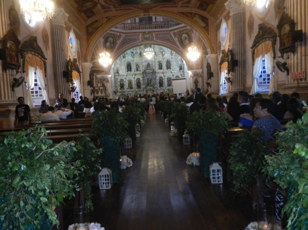 Eine Trauung in einer Kirche mit Gästen auf Bänken sitzend und im Gang stehend, dekoriert mit Zimmerpflanzen, Skulpturen, Vorhängen an den Fenstern und Kronleuchtern.