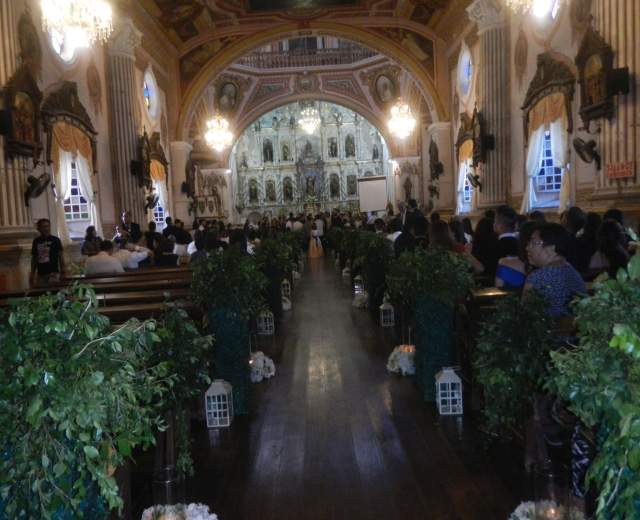 Eine Trauung in einer Kirche mit Gästen auf Bänken sitzend und im Gang stehend, dekoriert mit Zimmerpflanzen, Skulpturen, Vorhängen an den Fenstern und Kronleuchtern.