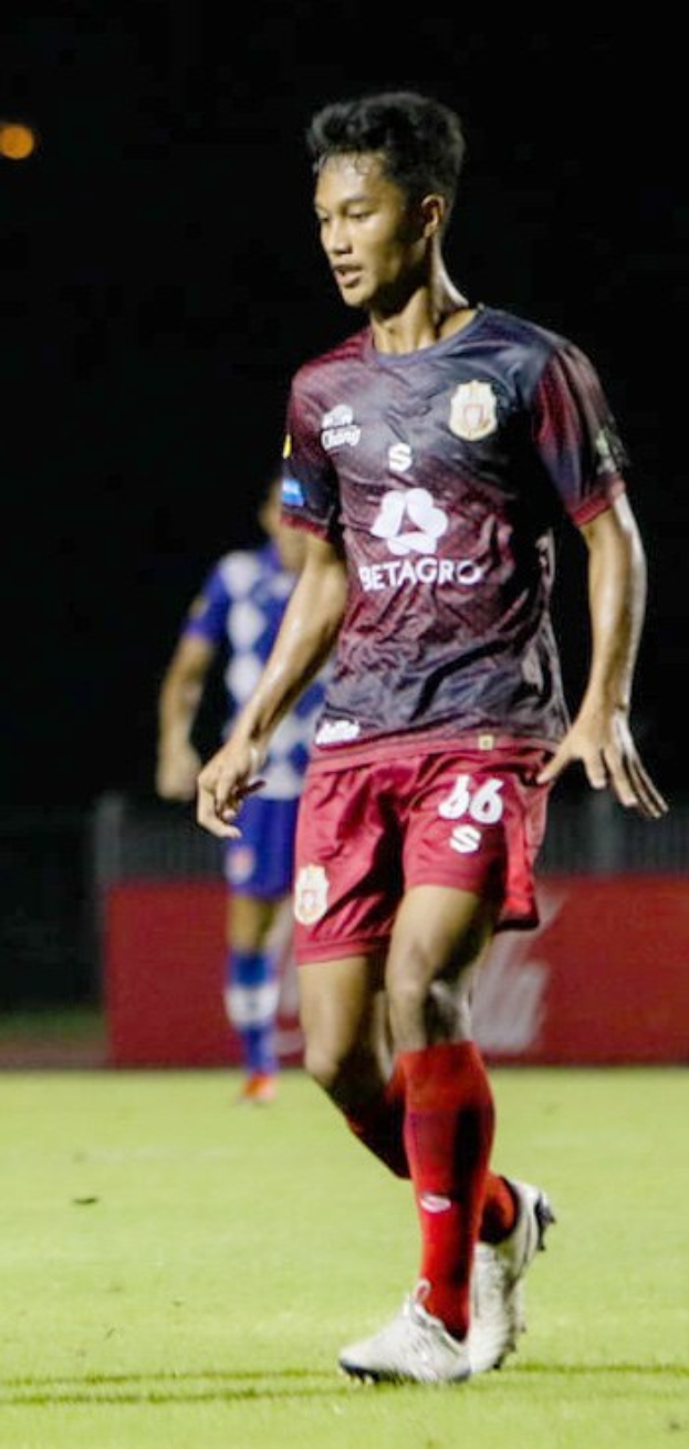 Ein professioneller Fußballspieler in einem marineblauen T-Shirt und roten Shorts, der einen Ball auf einem Feld kickt, mit einer anderen Person im Hintergrund und einer Tafel mit Text.