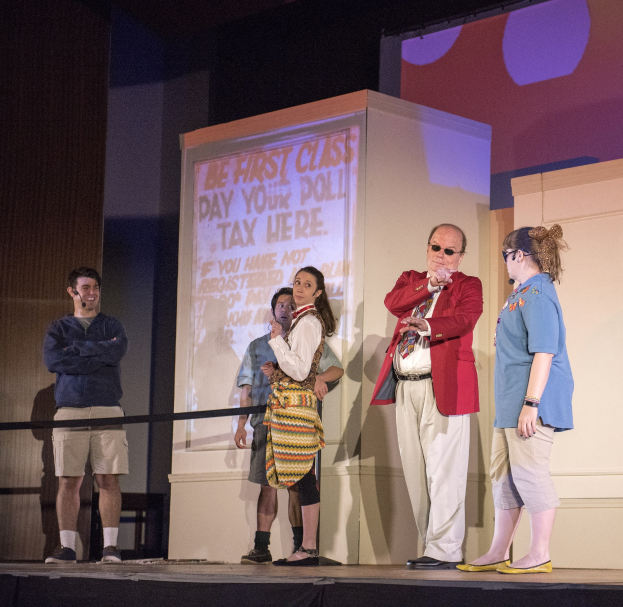 Eine Gruppe von Menschen auf einer Bühne mit einem Mann, der ein Mikrofon in der Mitte hält, ein Display hinter ihnen zeigt "The First Class Day Your Poll Tax Here", ein Zaun, eine Wand und Deckenleuchten.