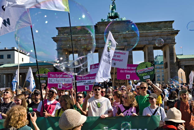 Menschenmenge vor dem Brandenburger Tor in Berlin, viele mit Mützen und Brillen, einige mit Fahnen und Schildern mit Text, mit Gebäuden, einem Bogen mit einer Statue darauf und einem Himmel voller Seifenblasen im Hintergrund.