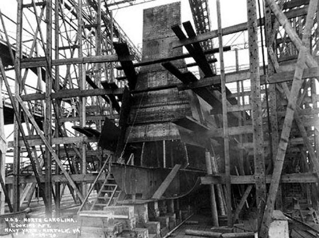Schwarzes und weißes Foto eines Schiffs in einem Schiffbau, mit Holzpfählen und Treppen im Vordergrund und Himmel im Hintergrund; Text "Marinebewaffnung" in der linken unteren Ecke.