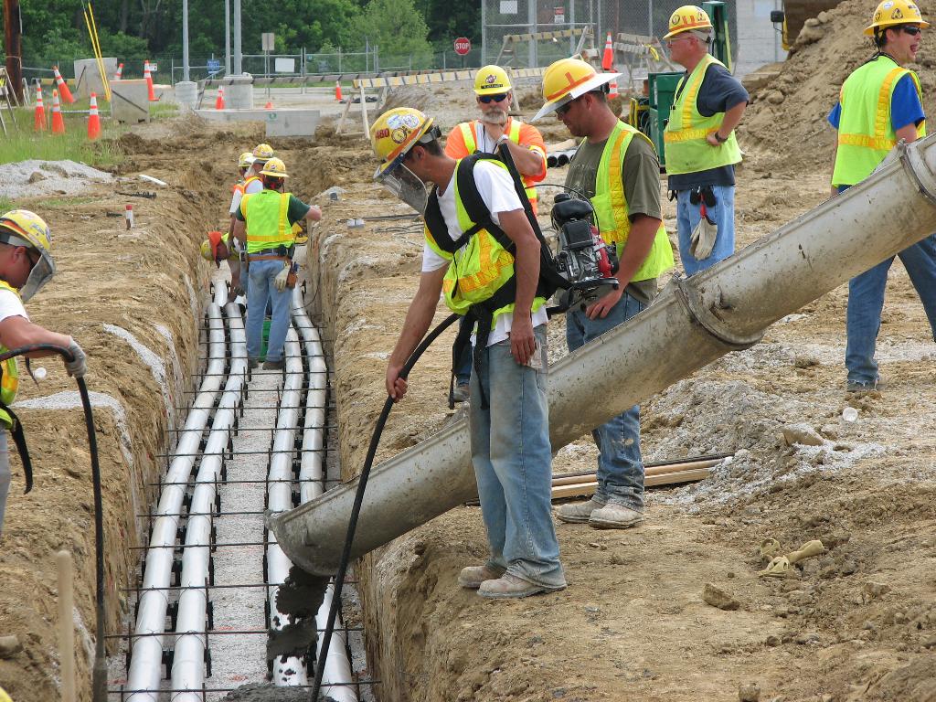Eine Baustelle mit Arbeitern in Westen und Helmen, Absperrbaken, Zaun, Bäumen, Pfählen, Erde und Rohren im Hintergrund.