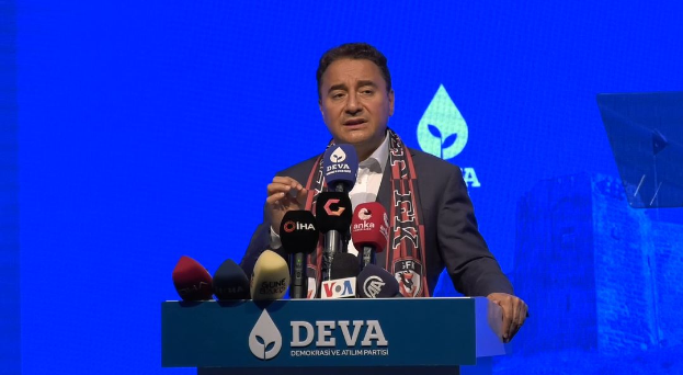 Ein Mann in einem Mantel und Schal steht an einem Podium mit Mikrofonen, ein Schild mit der Aufschrift "deva-Geschäftsführer sagt, es sei keine gute Idee für die Zukunft", ein Objekt und ein Bildschirm im Hintergrund.