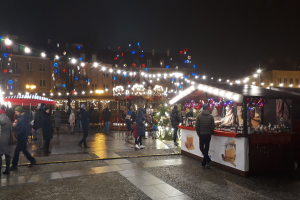 Ein belebter Weihnachtsmarkt bei Nacht in einer Stadt, mit Menschen, die zwischen geschmückten Ständen, Gebäuden, Bäumen und festlichen Lichtern im Hintergrund gehen.