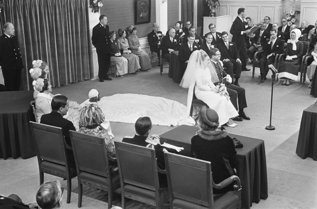 Schwarz-weiß-Foto einer Hochzeitszeremonie mit Menschen, die sitzen und stehen, einem Mann mit Mikrofon, einer Frau mit Blumenstrauß, einem Podium mit Mikrofon, Blumenschmuck, einem Vorhang, einem gerahmten Foto an der Wand und einer Tür im Hintergrund.