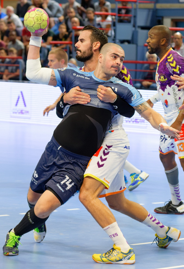 Männer spielen Hallenhandball in einer Arena, einer hält den Ball, mit Zuschauern im Hintergrund.
