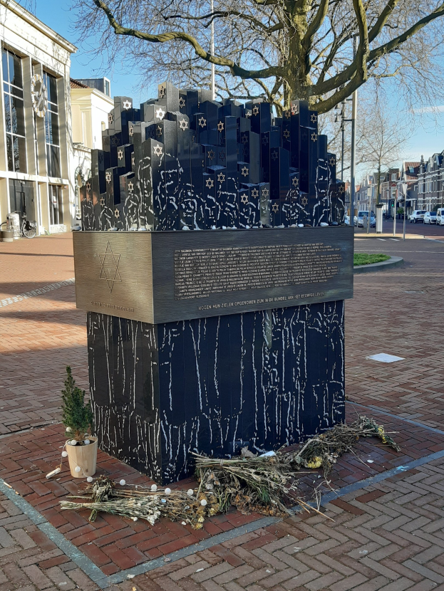 Denkmal in einem Stadtplatz gewidmet an Holocaust-Opfern, mit einem Baum davor, einer Topfpflanze und trockenen Blättern auf dem Boden, umgeben von Gebäuden, Fahrzeugen, Pfählen, Bäumen und Himmel.