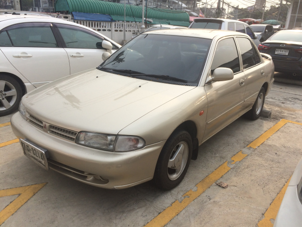 Ein goldfarbener Wagen auf einem Parkplatz neben anderen Fahrzeugen geparkt, mit Gebäuden, Strommasten, Drähten, Bäumen und einem klaren Himmel im Hintergrund.