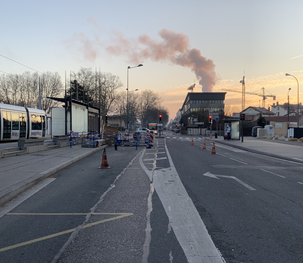 Eine Stadtstraße mit Verkehrskegeln, einem Bus, Laternen, Bäumen, Gebäuden, Kränen und Rauch, der aus einem entfernten Gebäude aufsteigt, vor einem Himmel.