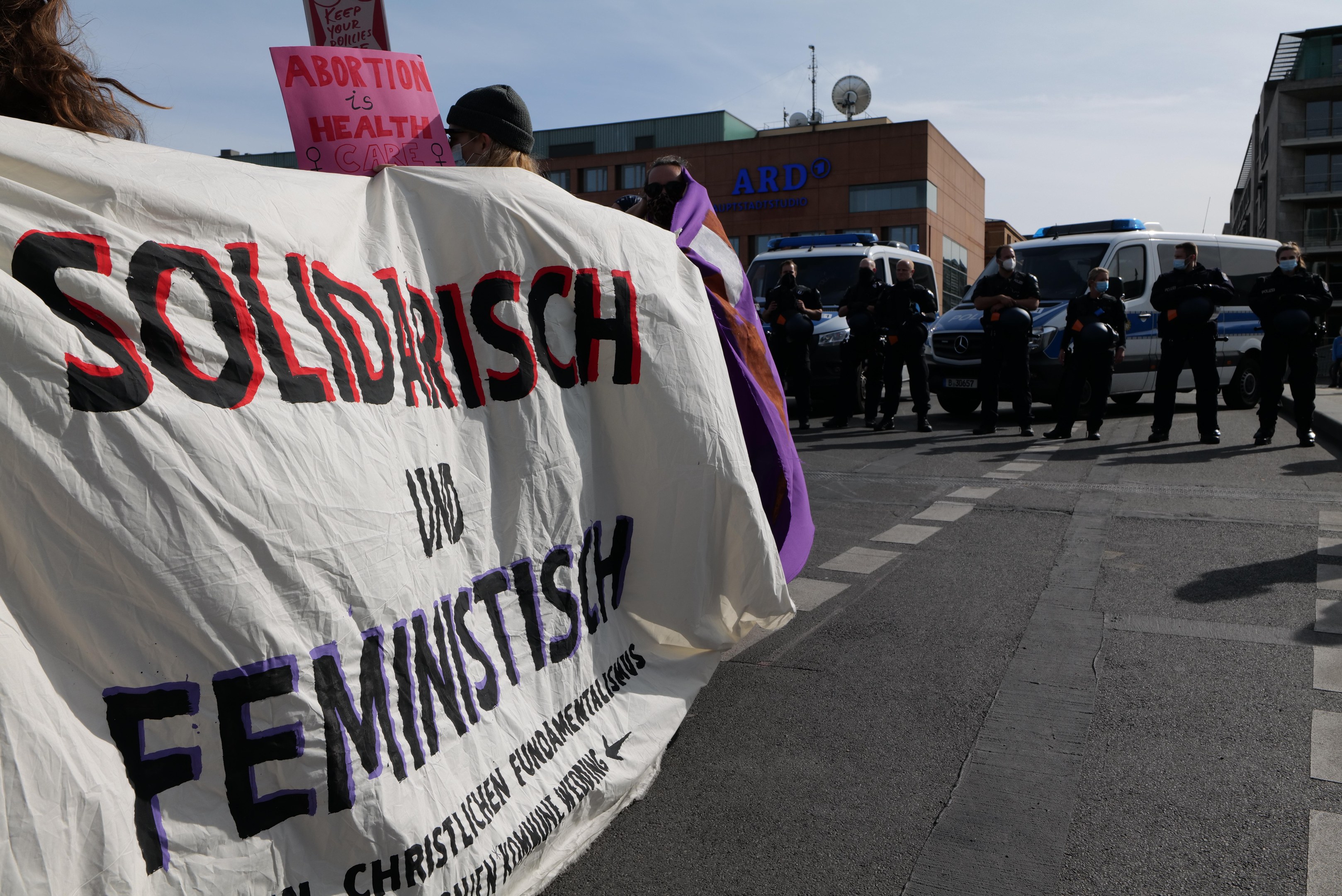 Eine Gruppe von Menschen marschiert auf einer Straße, hält ein Banner mit der Aufschrift "Solidarität und Feminismus" und hat parkende Fahrzeuge auf der Seite und Gebäude im Hintergrund unter einem klaren blauen Himmel.