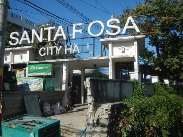Eingang zum Santa Rosa Rathaus mit einem Tor mit Text, Strommast mit Drähten, einer Gruppe von Menschen, Taschen, einem Behälter, Pflanzen, einer Wand, Bäumen und einem bewölkten Himmel.