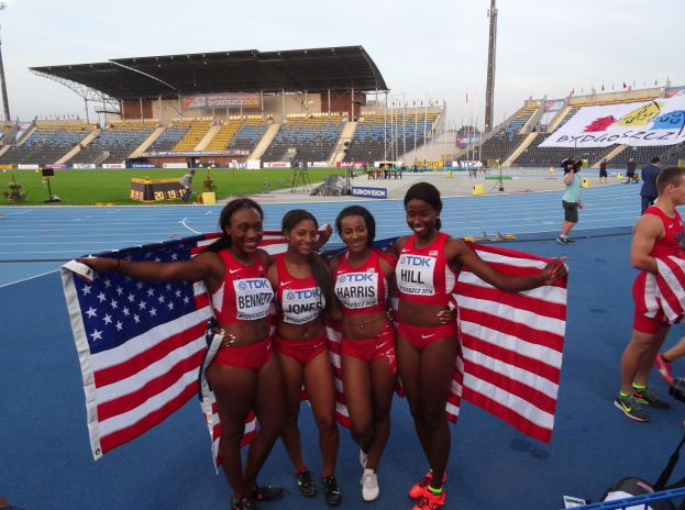 Vier Frauen in Sportkleidung stehen auf einer Laufbahn, lächeln und halten US-Flaggen, mit einem Stadion und Himmel im Hintergrund.