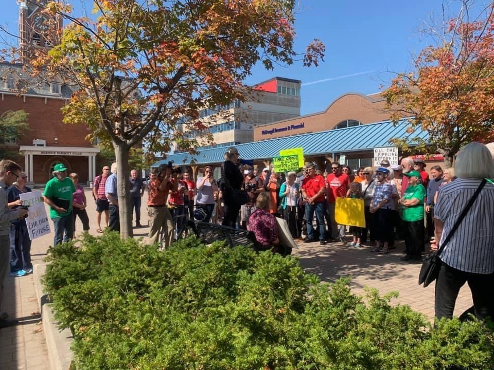 Eine Gruppe von Studenten demonstriert auf dem Campus der University of Michigan und hält Schilder hoch, einige tragen Mützen und tragen Taschen oder Kameras, mit Bäumen und Himmel im Hintergrund.