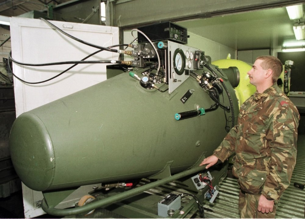 Ein Mann in militärischer Uniform steht neben einer großen grünen Bombe in einem unordentlichen industriellen Setting mit Maschinen und beleuchteten Wänden, mit Text unten.