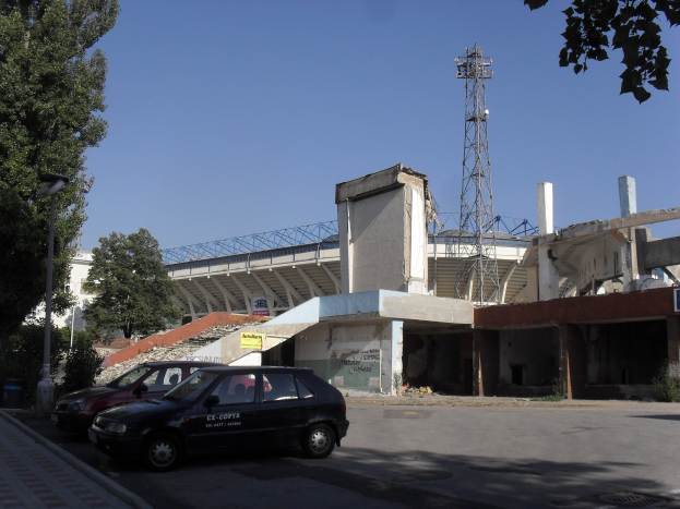 Alter Stadion mit parkenden Autos davor, umgeben von Gebäuden, Bäumen, Polen und einem Turm unter einem sichtbaren Himmel.