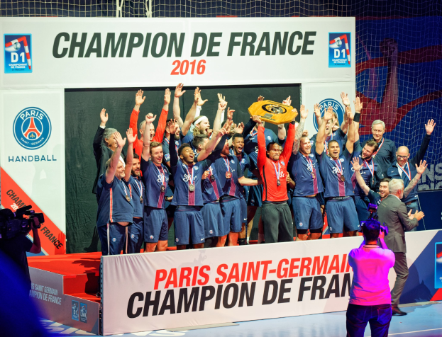 Gruppe von Männern, die einen Pokal auf der Bühne halten, mit einer Videokamera, die aufnimmt, und einem "Champion de France 2016"-Schild im Hintergrund.