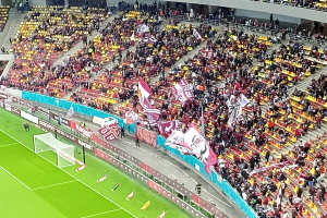 Ein großes Stadion voller Zuschauer bei einem Fußballspiel, mit einem Tor mit Netz auf der linken Seite, einigen Menschen, die stehen und auf Stühlen sitzen, mehreren Schildern und im Hintergrund Treppen, Geländer und eine Wand.