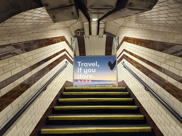 Eine Treppe in einer U-Bahn-Station mit einem Schild, das oben 'Reise, wenn du wagst' trägt, flankiert von Geländern und sichtbaren Stationswänden im Hintergrund.