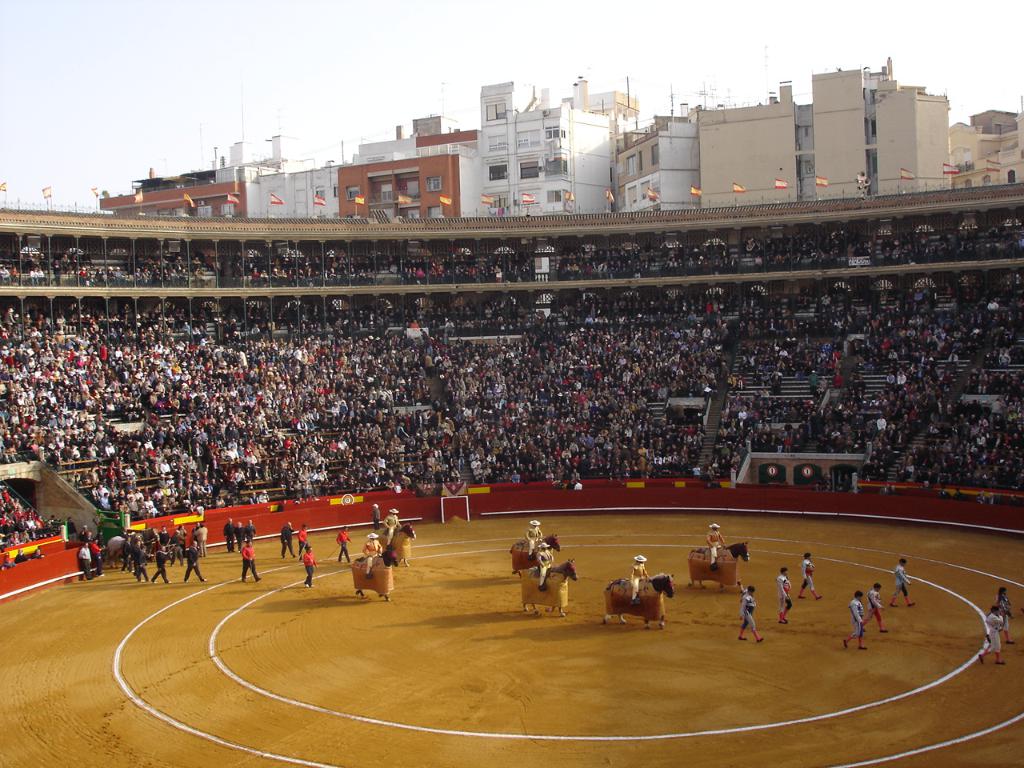 Eine Menschenmenge, die vor einem Stadion voller Zuschauer spazieren geht und reitet, mit Fahnen, Gebäuden und dem Himmel im Hintergrund.