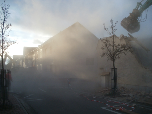 Ein Gebäude wird mit Rauch aus dem Schornstein abgerissen, ein Kran im Hintergrund, Menschen auf dem Boden und ein bewölkter Himmel.