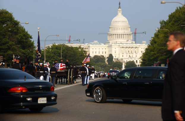 Trauerzug mit Menschen in Mützen und Fahnen vor dem Kapitolsgebäude, begleitet von Fahrzeugen und Bäumen unter einem klaren Himmel.