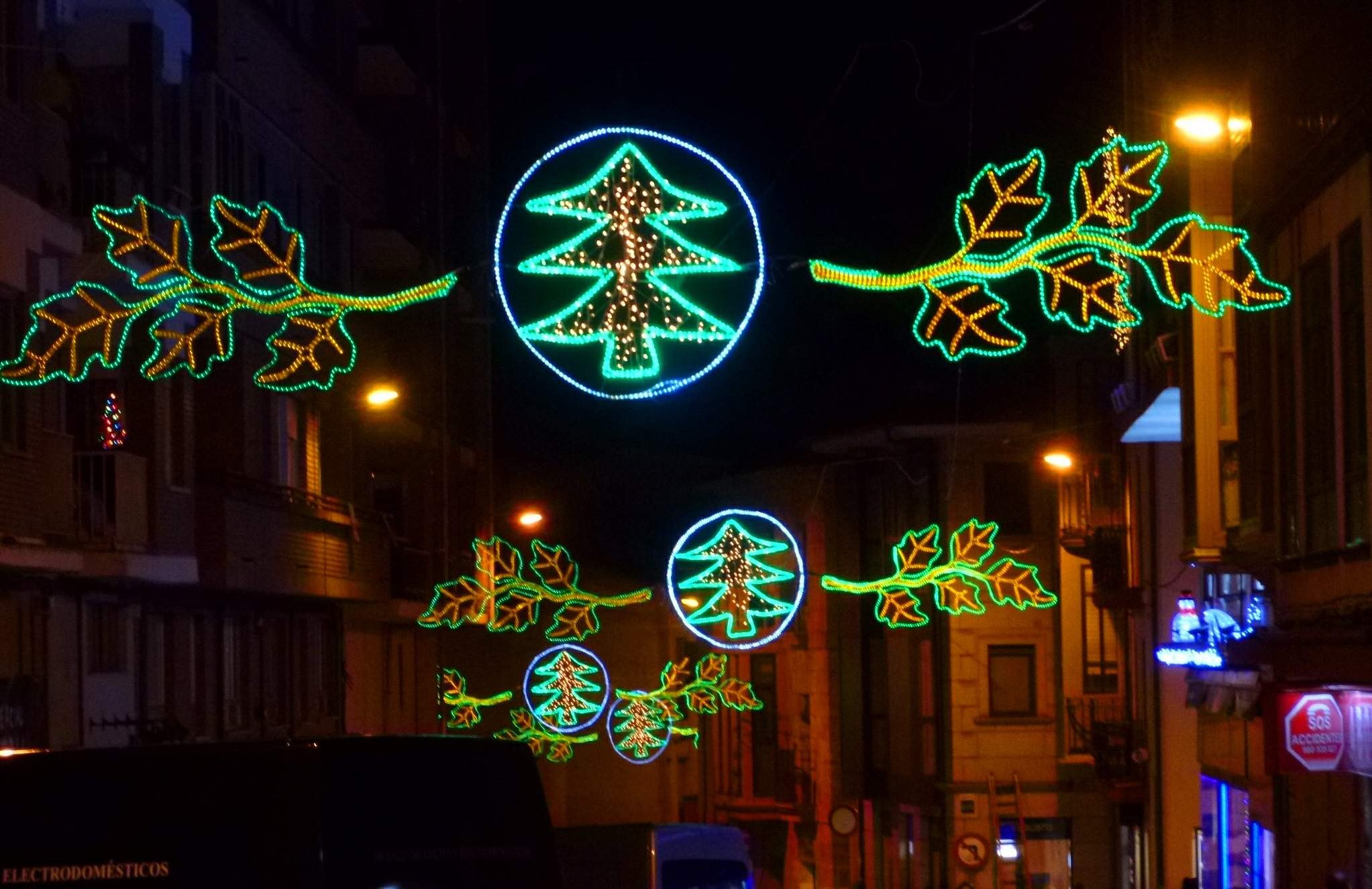 Eine städtische Straße, geschmückt mit festlichen Weihnachtslichtern, die Blätter und andere Dekorationen zeigen, mit Fahrzeugen, die an Gebäuden auf beiden Seiten vorbeifahren.