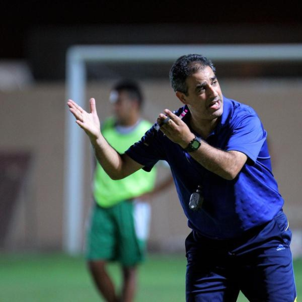 Ein Mann in einem blauen T-Shirt und Hose steht auf einem Fußballfeld und hält ein Mikrofon in der Hand, während er auf Spieler zeigt; im Hintergrund ist eine weitere Person in einem grünen Kleid neben einem Torpfosten und einer Wand zu sehen.
