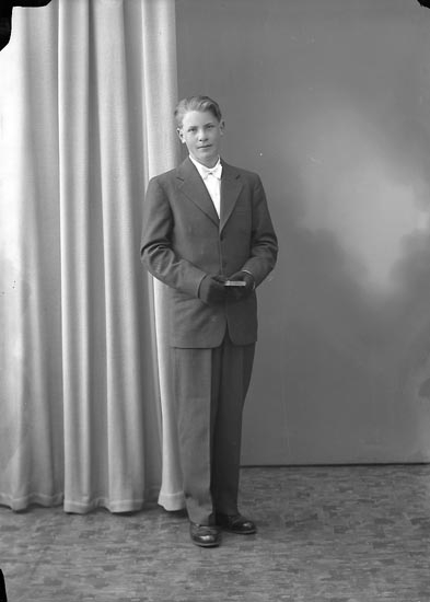 Ein Mann im Anzug mit einem weißen Hemd und einer gemusterten Krawatte steht vor einem Vorhang, sein Haar ordentlich frisiert und einen ernsten Gesichtsausdruck.