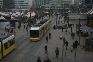 Eine belebte Stadtstraße mit zahlreichen Fußgängern, einige mit Schirmen, und Radfahrern, umgeben von Gebäuden, Zelten, Pfählen, Laternen und Bäumen unter einem klaren blauen Himmel.